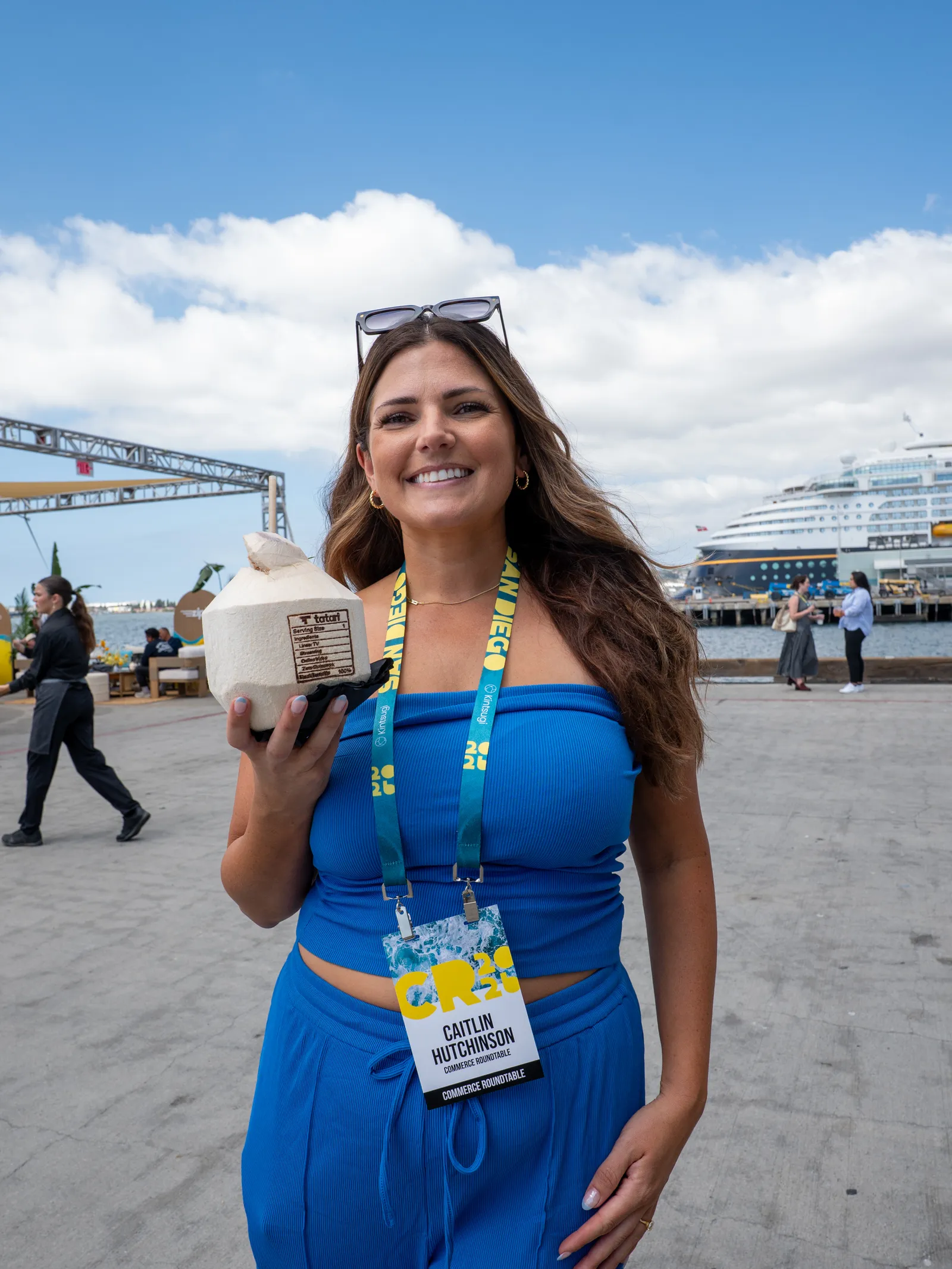A smiling attendee in a blue dress holds a coconut drink on the waterfront.