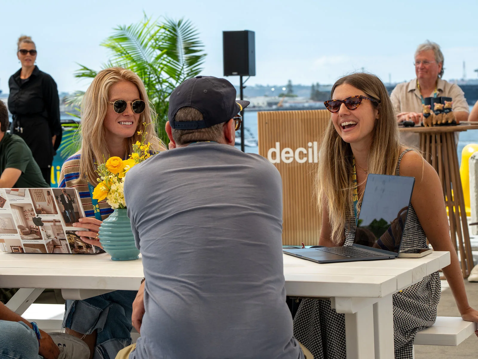 Attendees laugh together at a table under shade sails by the water.